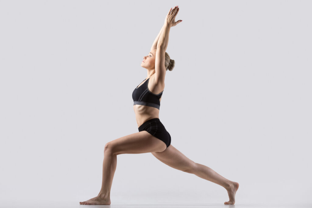 Postura del Guerrero I, pose Virabhadrasana One, ejercitándose con ropa deportiva negra, estudio de cuerpo entero, fondo gris.